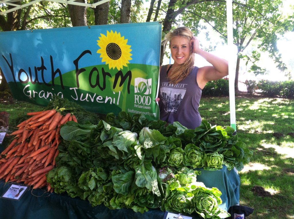 a young blond woman holds a youth farm sign behind bunches of fresh produce