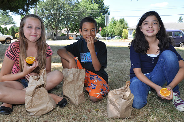 three kids sit on the ground outside smiling and eating lunch